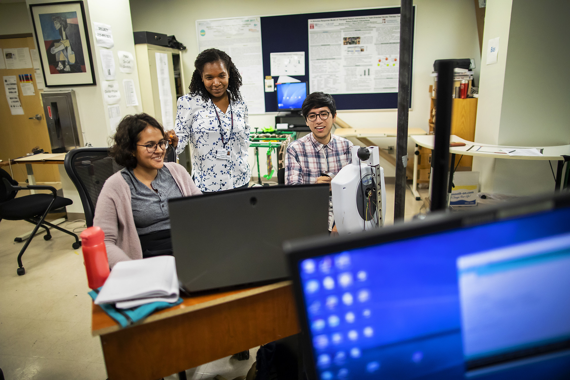 Two seated postdocs and a standing instructor with a lanyard look at a monitor screen in a computer lab.